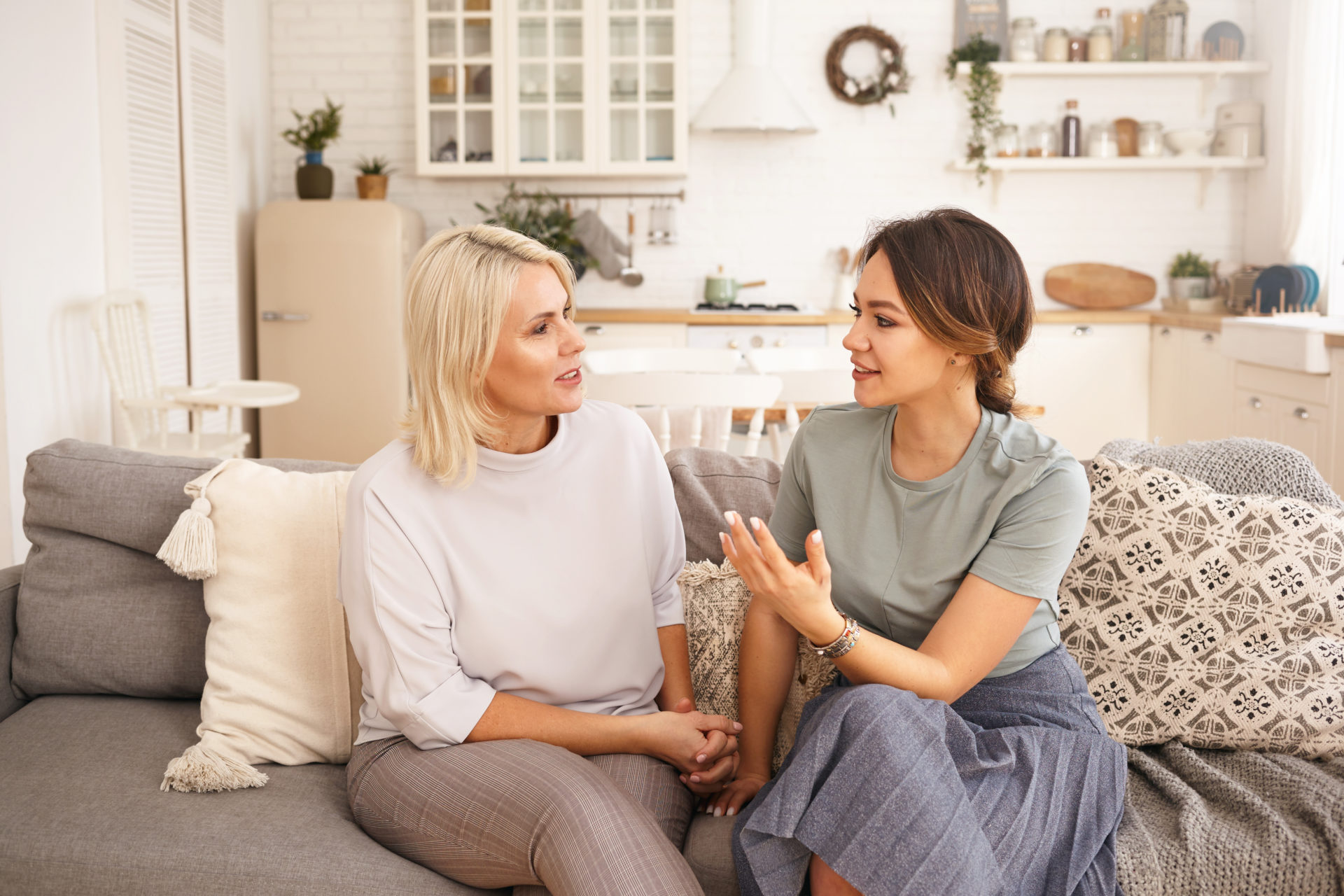 Two Caucasian women having nice lively conversation in living room. Cute young woman gesturing emotionally, proving something while talking to her middle aged mother on sofa in cozy interior