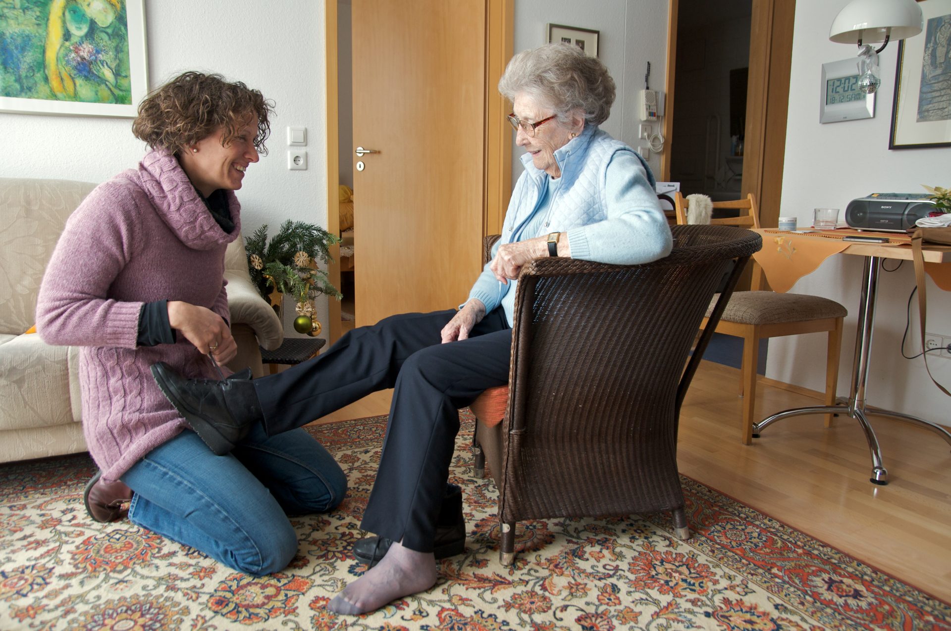 Elderly lady sitting in a chair receiving help putting her shoes on.